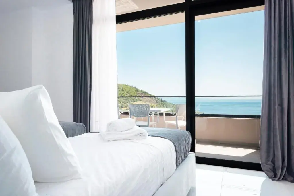 A bed in white linen with folded towels beside a full-height black-framed sliding door standing open onto a balcony with a table and chairs, the sea and a green headland beyond