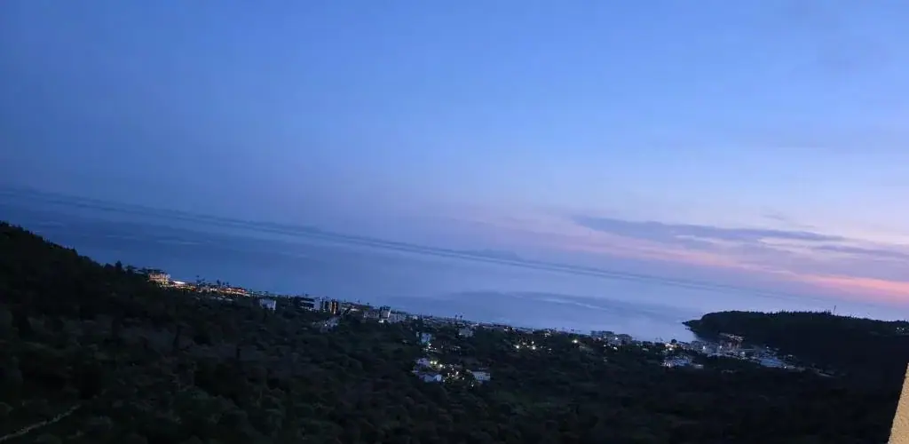 Dusk over the Himarë coast seen from a balcony at Wait 'n Sea: a dark wooded slope, the lights of the shore strung along the bay, and pink cloud over the sea