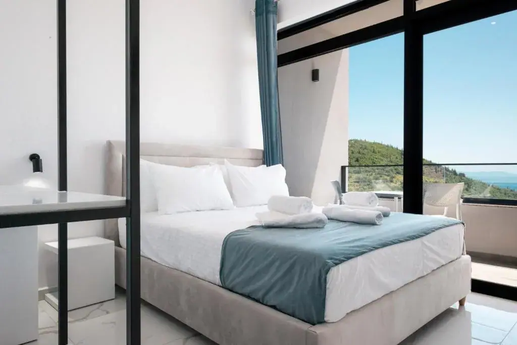 A bed with a teal throw and folded towels behind a black steel-framed partition, beside a glass wall onto a balcony with the wooded ridge and the sea behind it