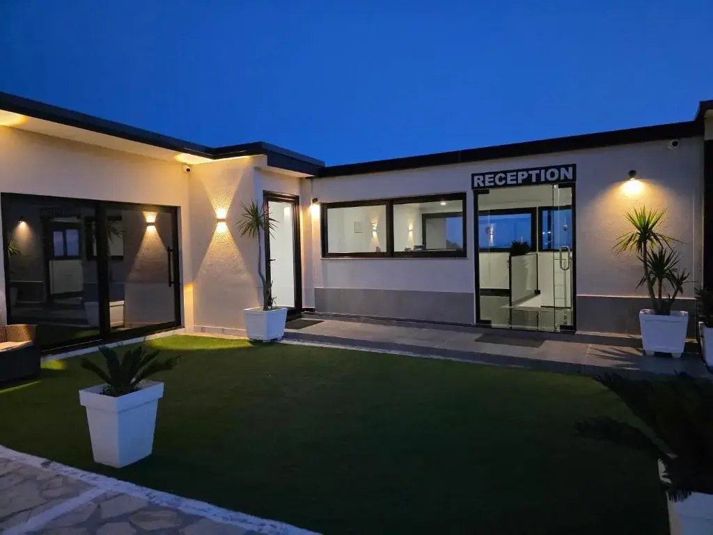 The single-storey white reception block at Wait 'n Sea lit by uplighters at dusk, its RECEPTION sign over black-framed glass, with a lawn and potted palms in front