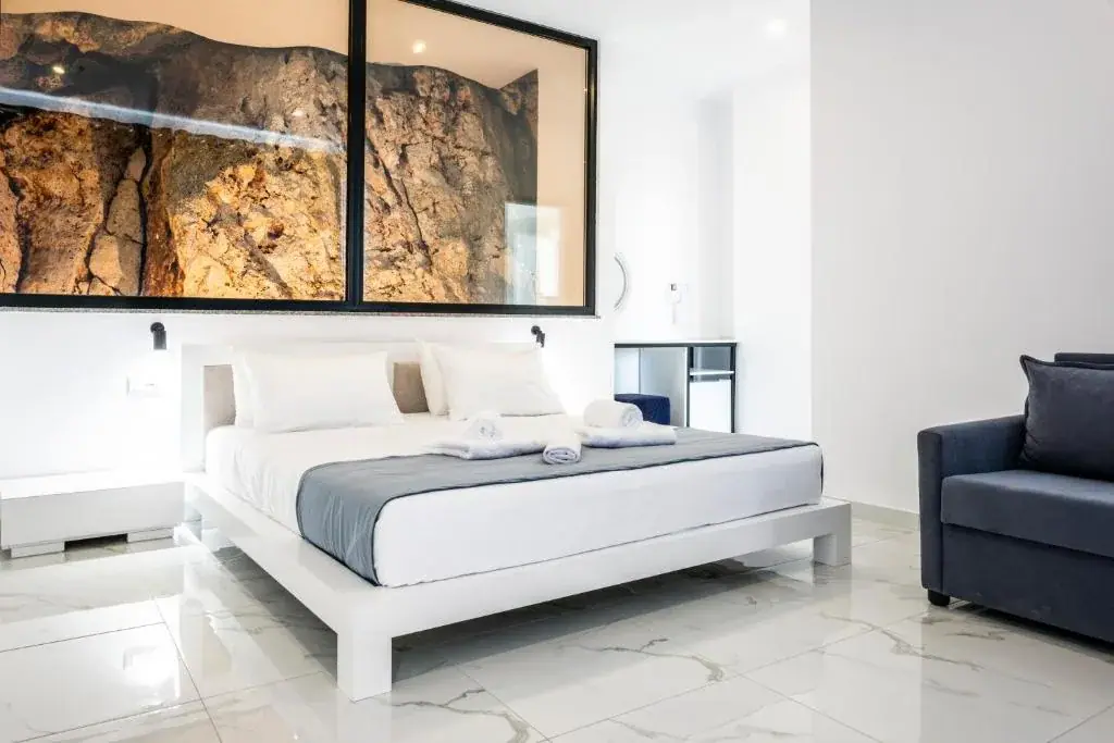 A low white platform bed on white marble with a blue sofa beside it, under a black-framed internal window onto the raw ochre rock face behind the building