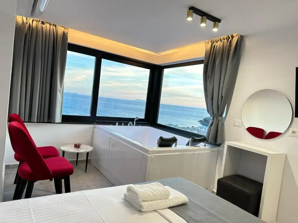 A white spa bath set beneath the corner windows of a room at Wait 'n Sea, with a red velvet chair, a round mirror and the sea and a rocky point outside