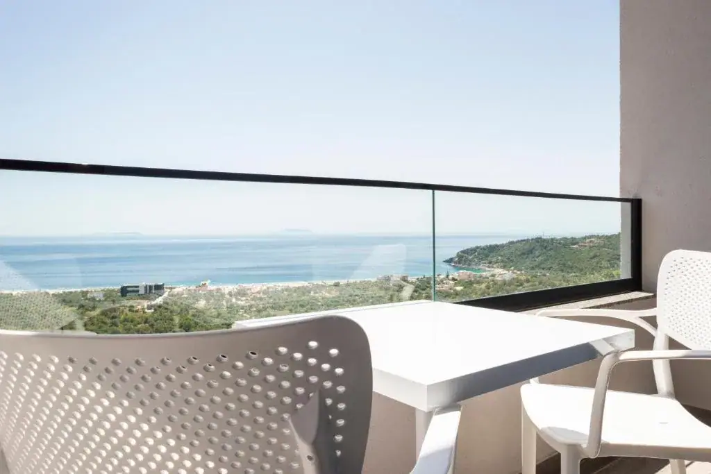 A white table and two chairs on a balcony at Wait 'n Sea above the whole bay, with a low island on the horizon beyond the glass balustrade