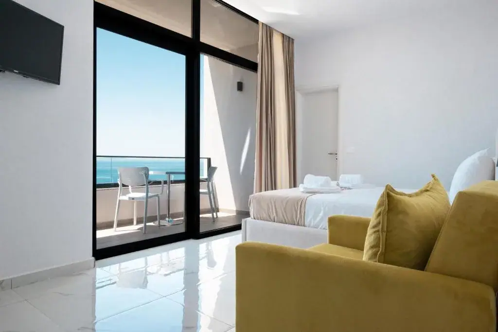 A room at Wait 'n Sea with a mustard velvet sofa, a bed with folded towels and a black-framed glass wall onto a balcony set with a table and chairs over the sea