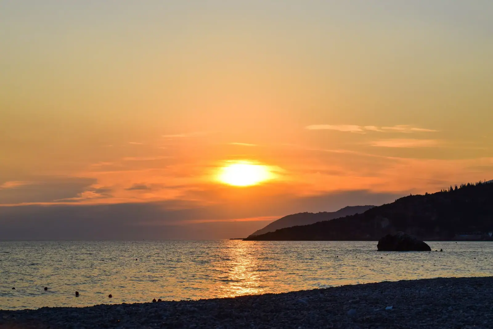 Livadhi Beach at dusk: the sun going down over open water, the headland off to one side, the shingle in the foreground.