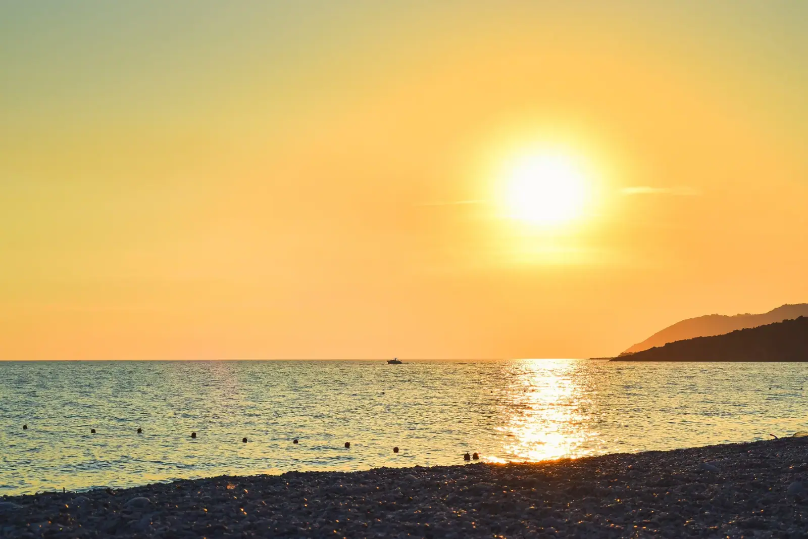 The sun dropping over Livadhi Beach, the swim-line buoys on flat water and the grey shingle in the foreground.