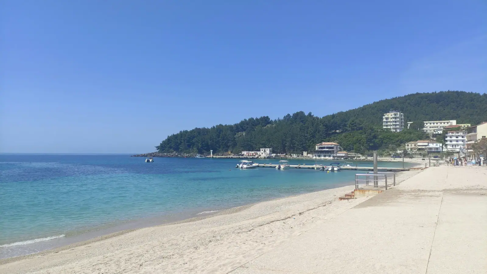 The shore at Himarë with the jetty and moored boats, the wooded headland behind and the town climbing the hill