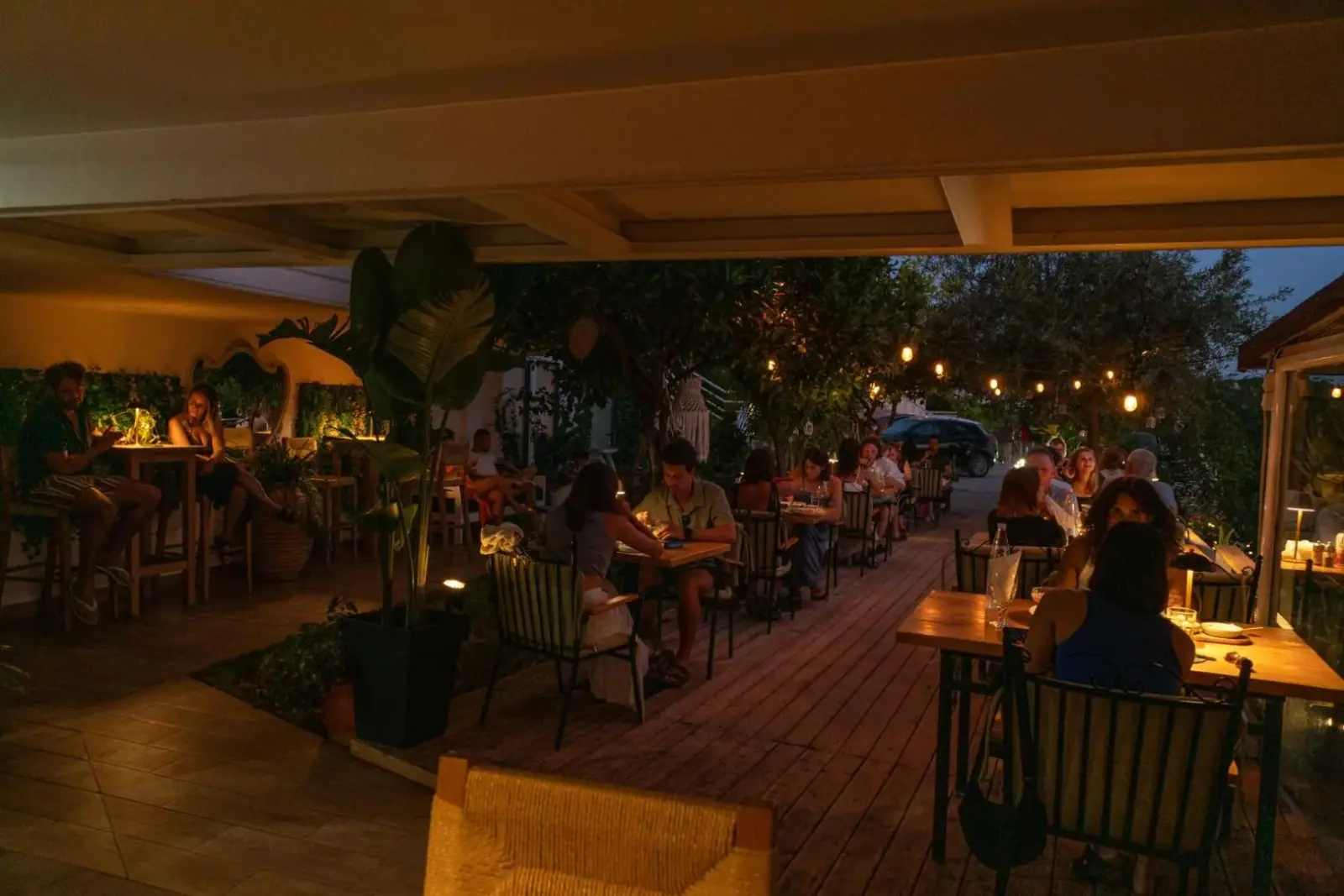A long restaurant deck after dark, strung with lights and planted with palms, every table taken