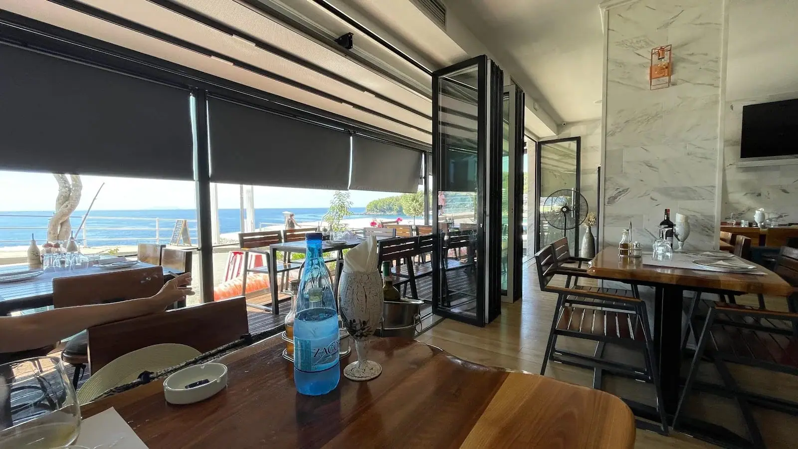 The dining room with marble walls and live-edge timber tables, its folding glass front open to the terrace and the sea