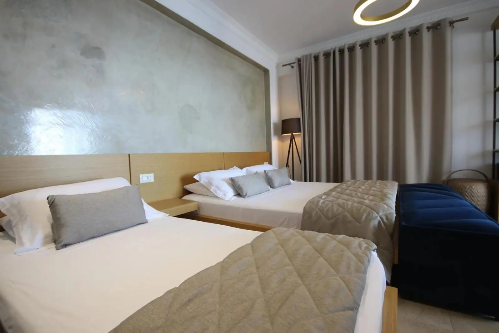 Two beds side by side under quilted throws, with timber headboards under a panel of polished plaster and a standing lamp