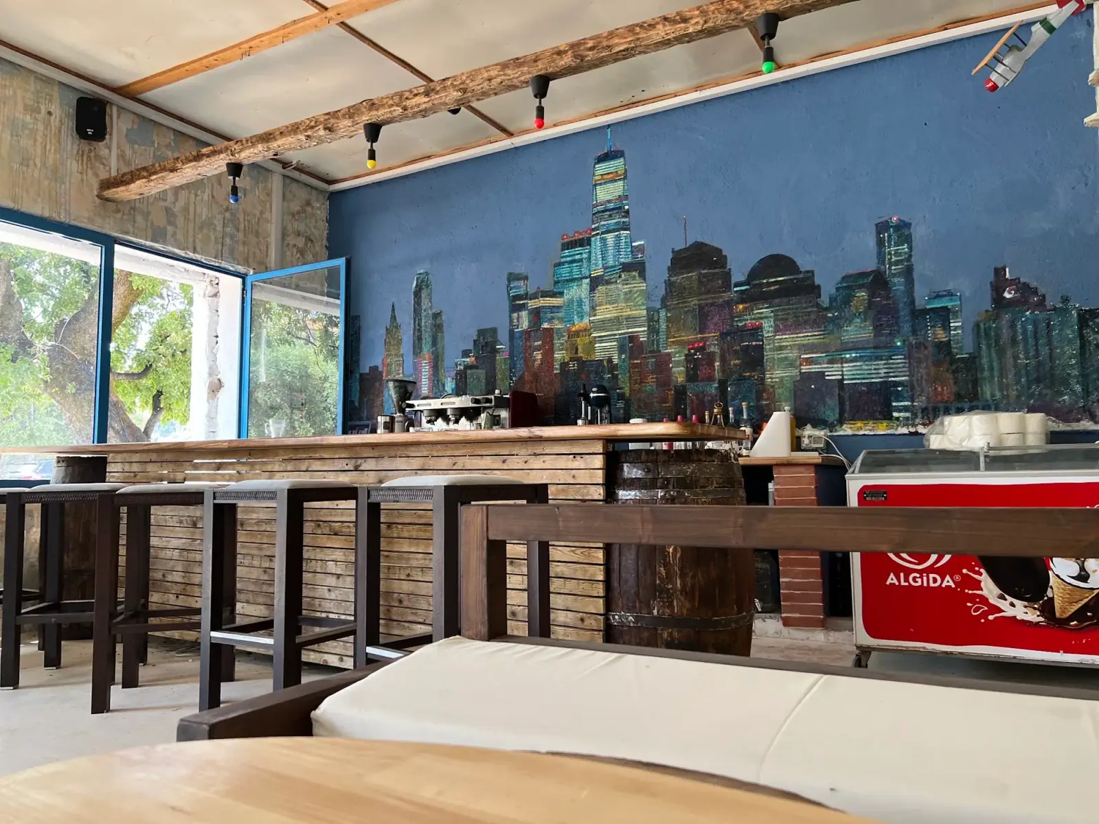 The bar counter at Bar 300 in front of a painted New York skyline at night, with bar stools and a barrel at the end of the slatted counter