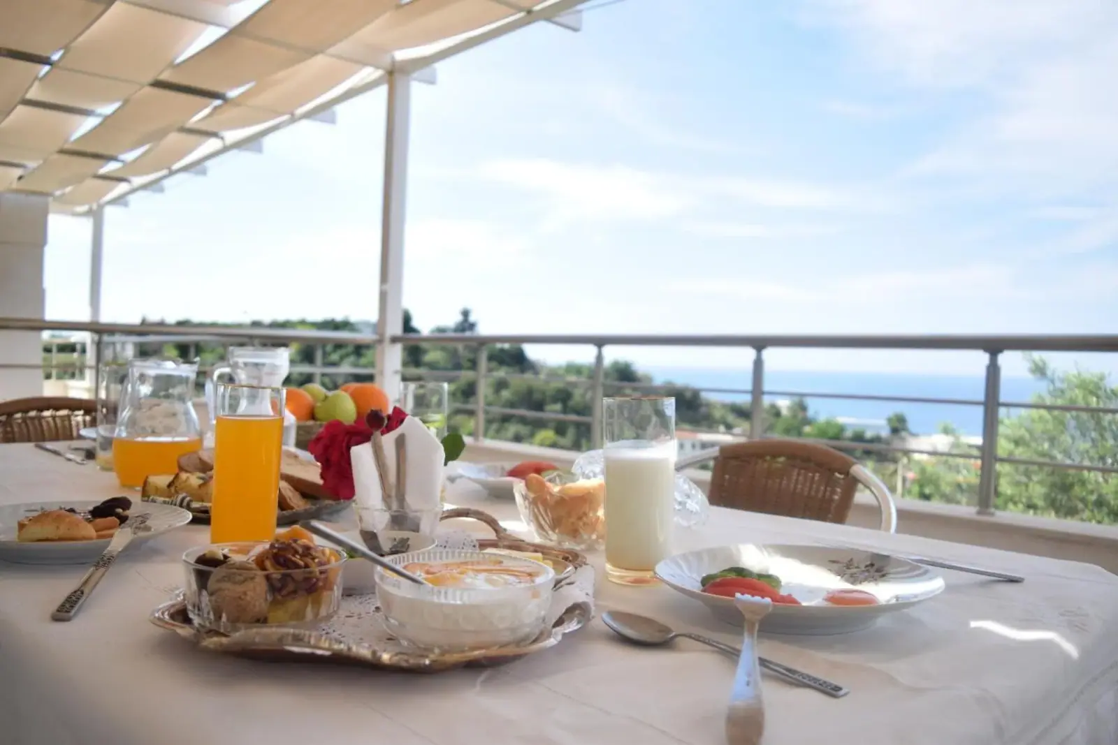 A table laid with jugs of juice, pastries and fruit on a covered terrace, the sea beyond the rail