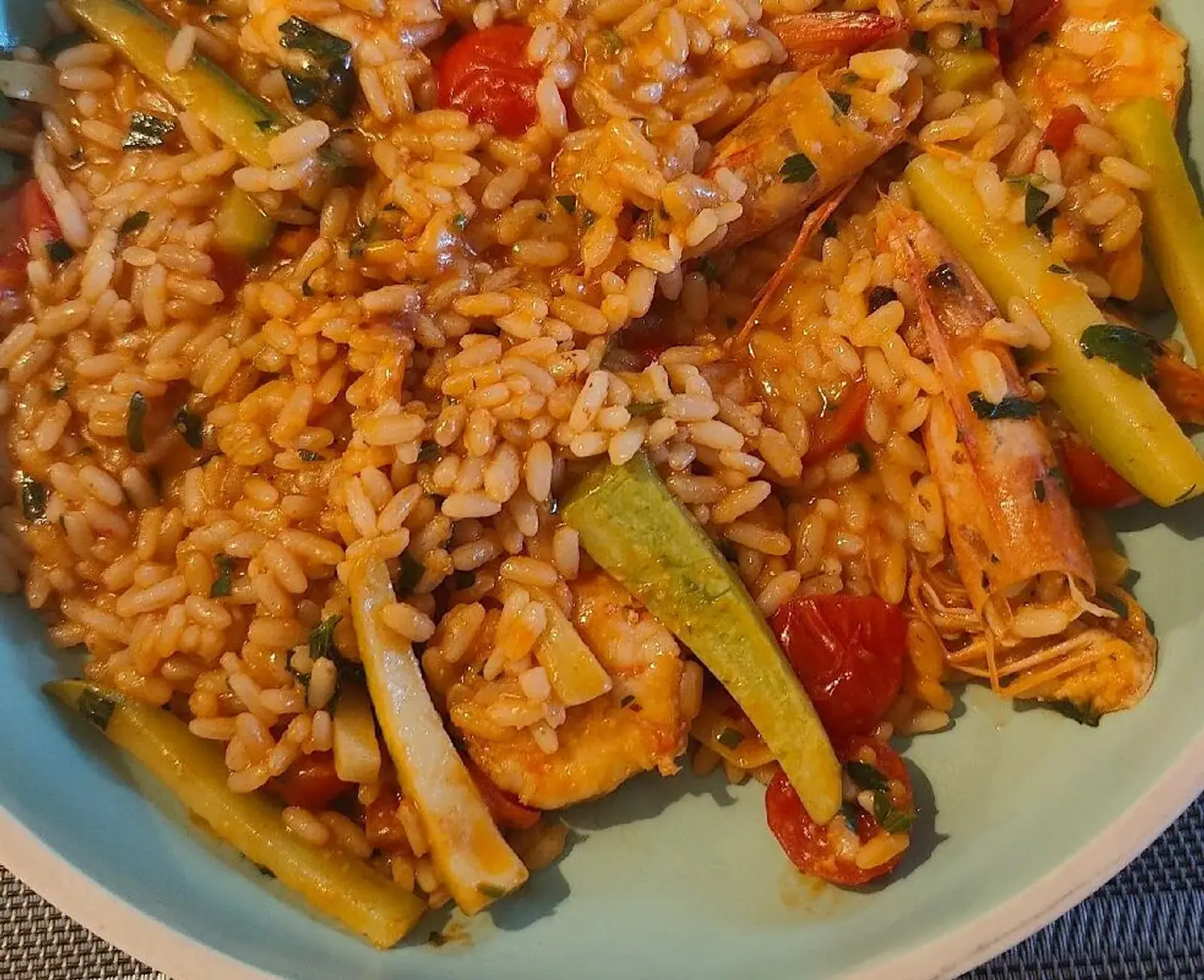 Close view of tomato-tinted rice with whole prawns, cherry tomatoes, courgette batons and chopped parsley, served in a pale celadon bowl