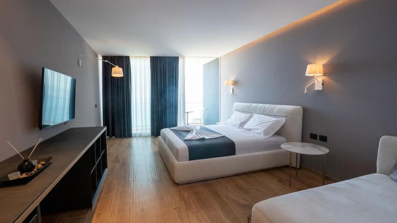 A guest room in pale grey with cove lighting, a white upholstered bed, petrol-blue curtains at a balcony door and a marble-topped side table on slim brass legs