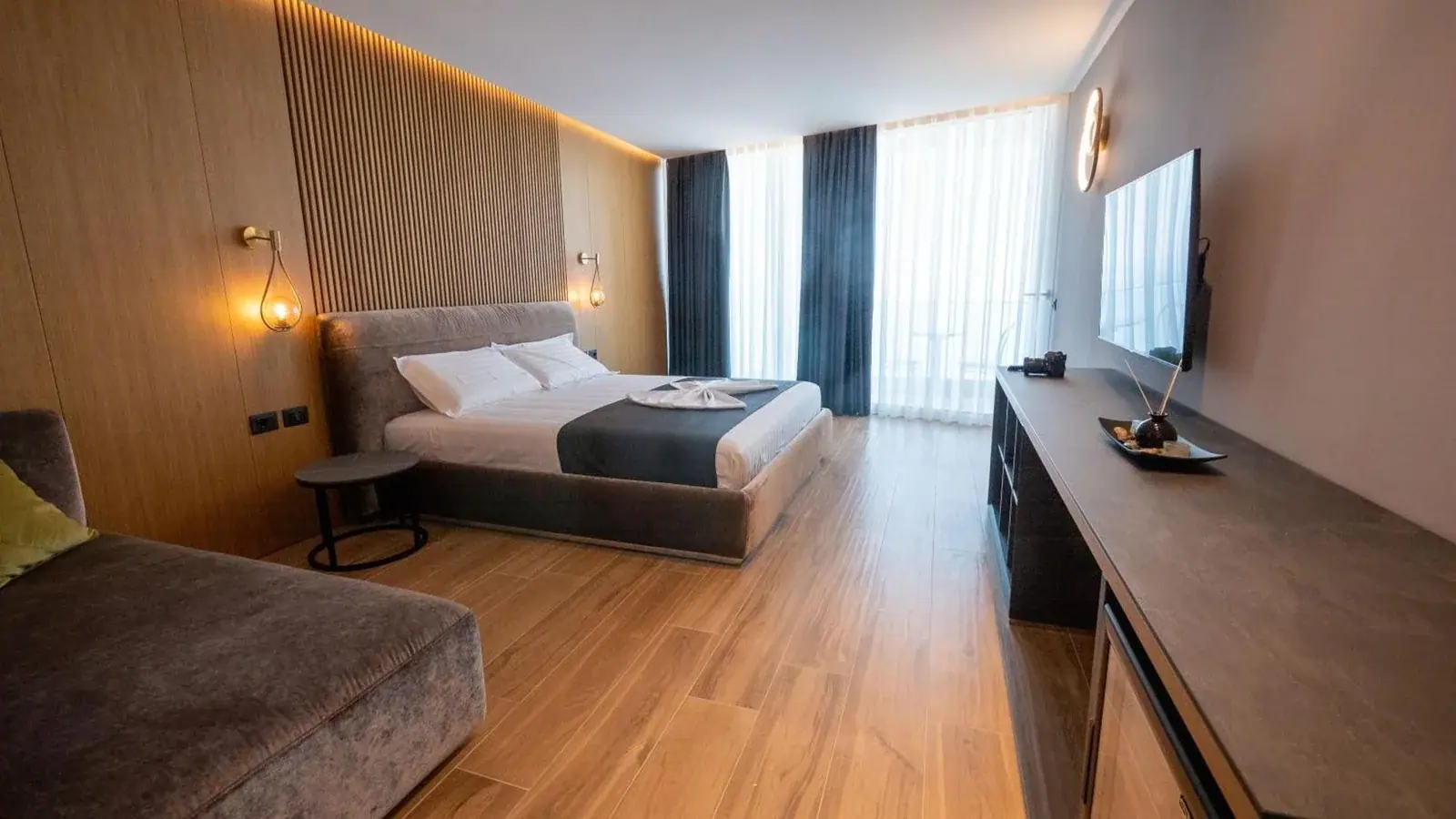 A guest room with an oak slatted wall lit from behind, brass globe reading lamps, a taupe upholstered bed with a charcoal runner, and full-height curtains drawn back from a balcony door