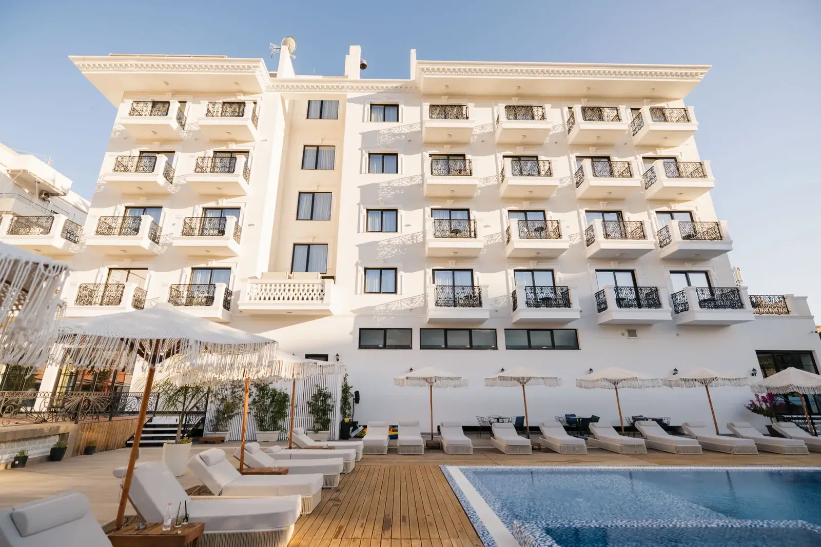 The rear elevation of Chimaera Hotel in Himarë: tiers of white balconies with black wrought-iron rails above the pool deck, its loungers and fringed umbrellas