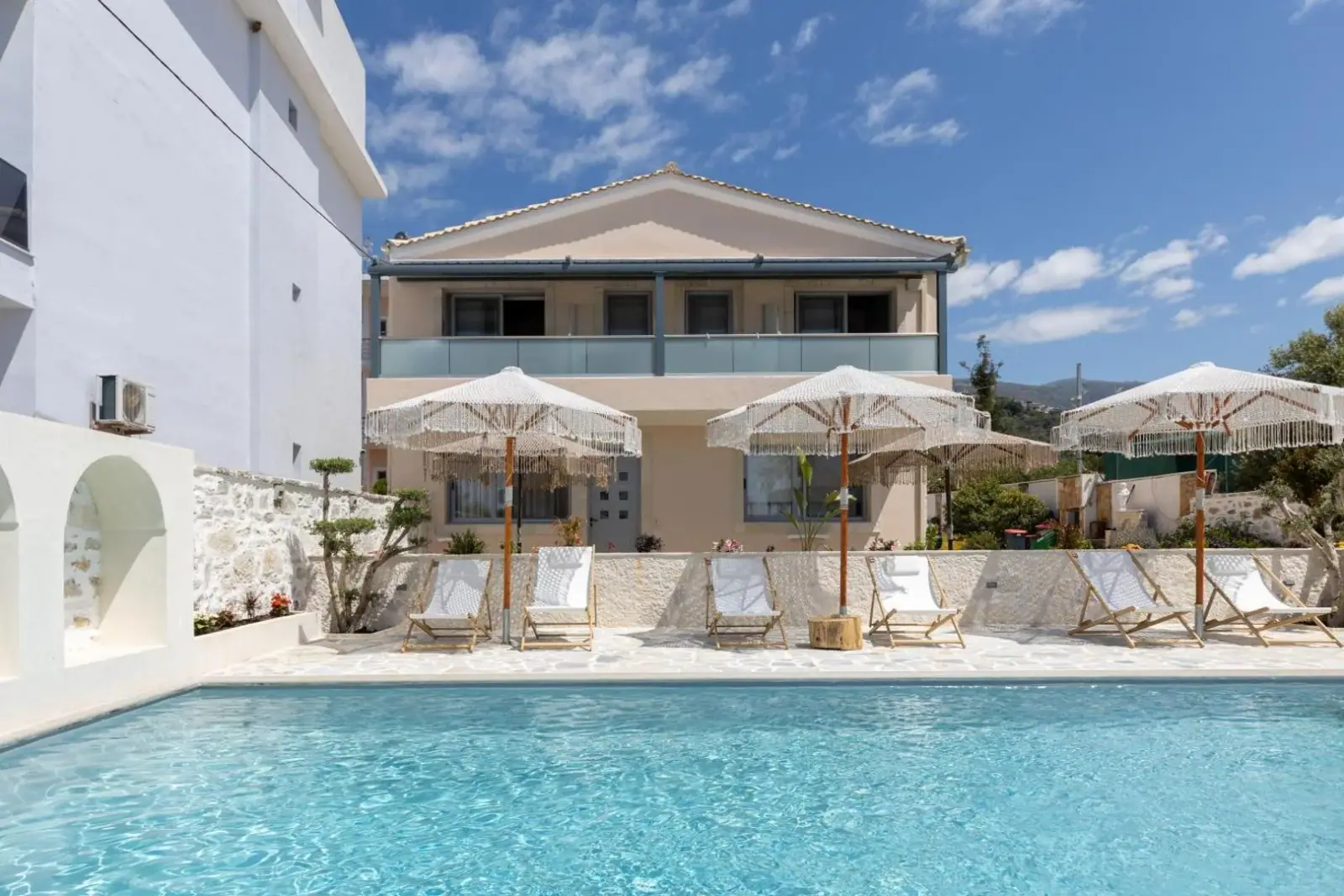 The pedimented front of Dimitri Hotel above its pool at Livadhi: fringed parasols and pale deck chairs along the stone coping, and a white arched niche in the wall at the left