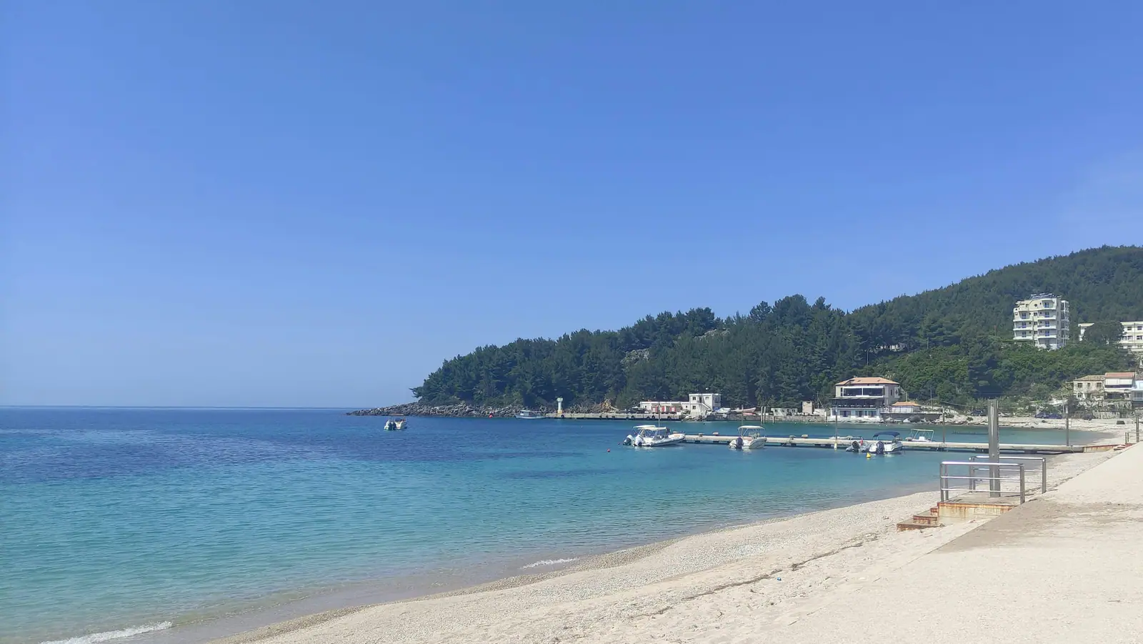 Spile beach at Himarë looking along the bay to the jetty, its moored boats and the pine-covered headland beyond