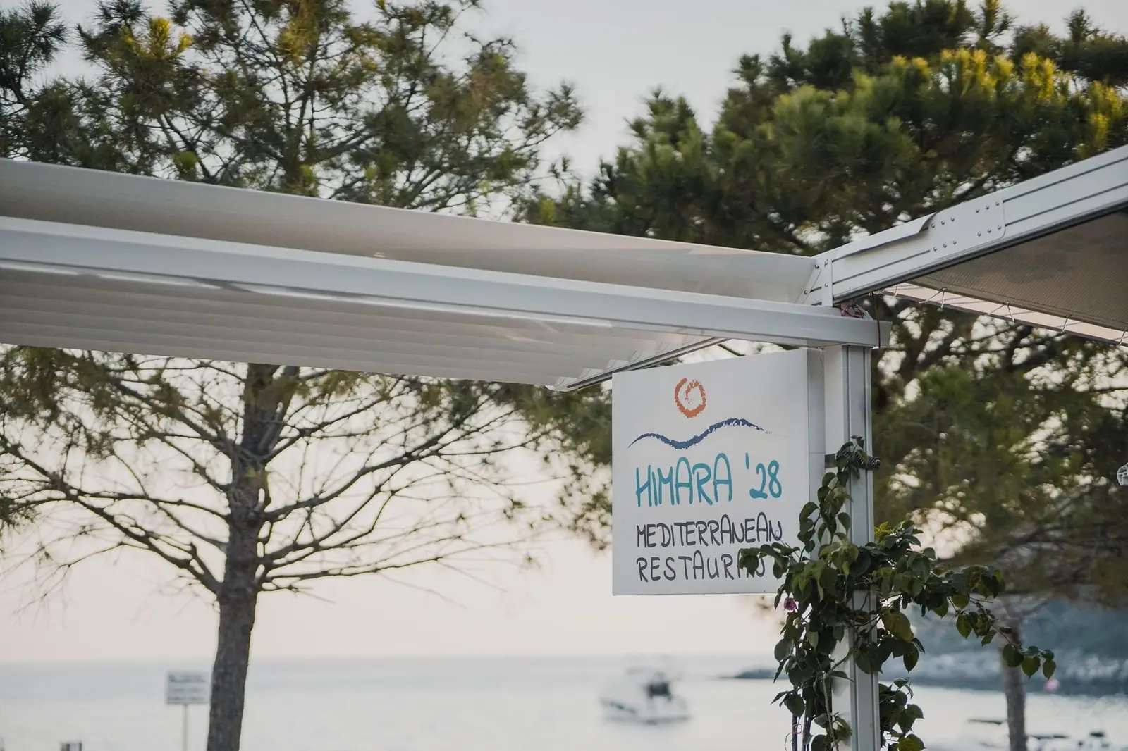 A hand-painted board hung from the awning frame, a drawn sun and wave above the lettering Himara '28 Mediterranean Restaurant, the sea and pines behind