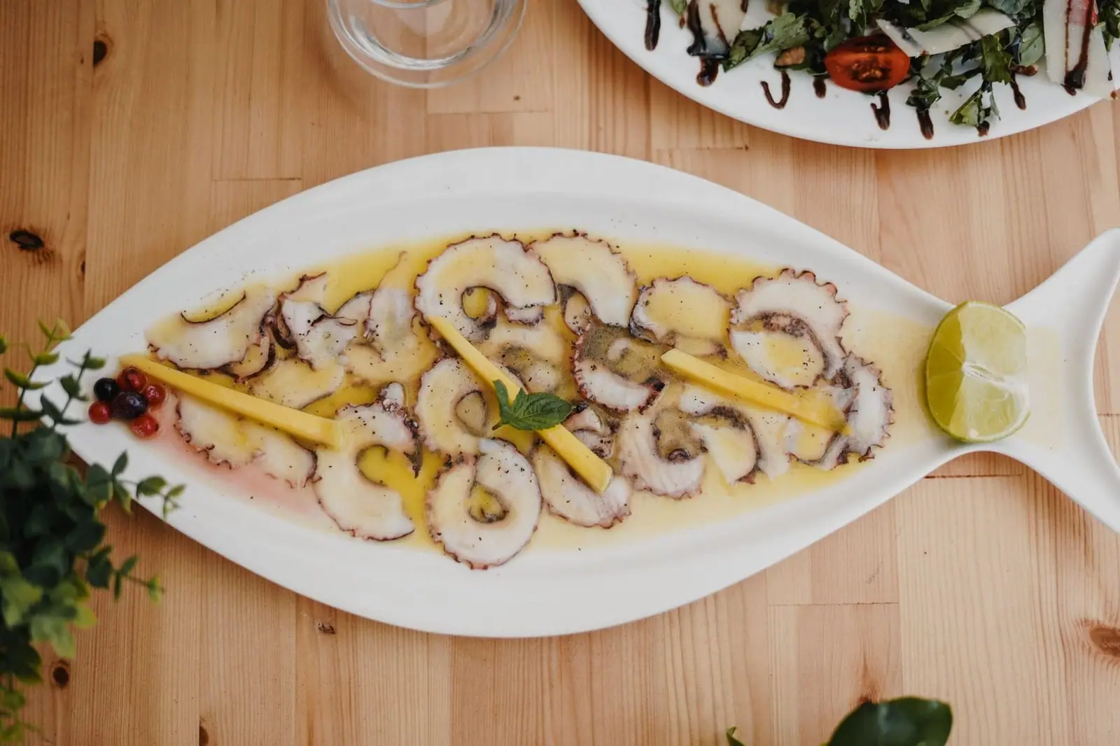 Thinly sliced octopus in oil and lemon along a white fish-shaped dish, with lime at the tail