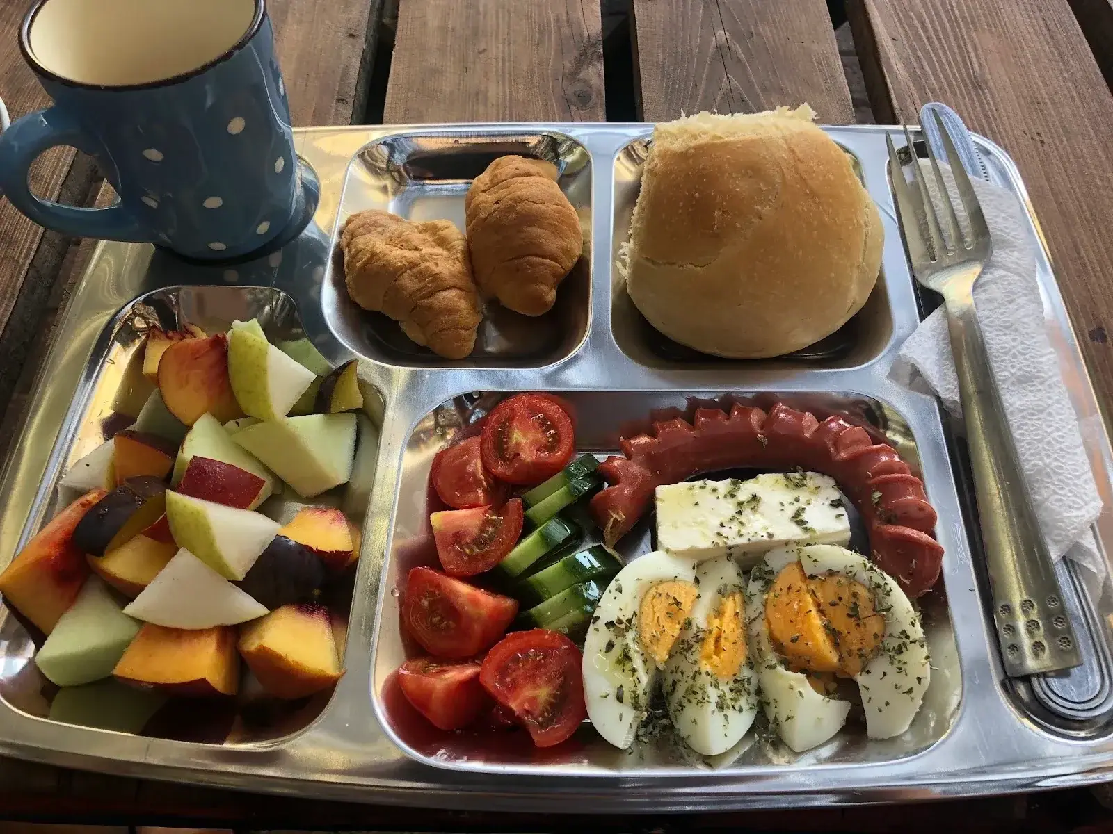 The free breakfast at Himara Downtown Hostel on a steel compartment tray: cut fruit, croissants, a bread roll, tomato, cucumber, boiled eggs, cheese and sausage