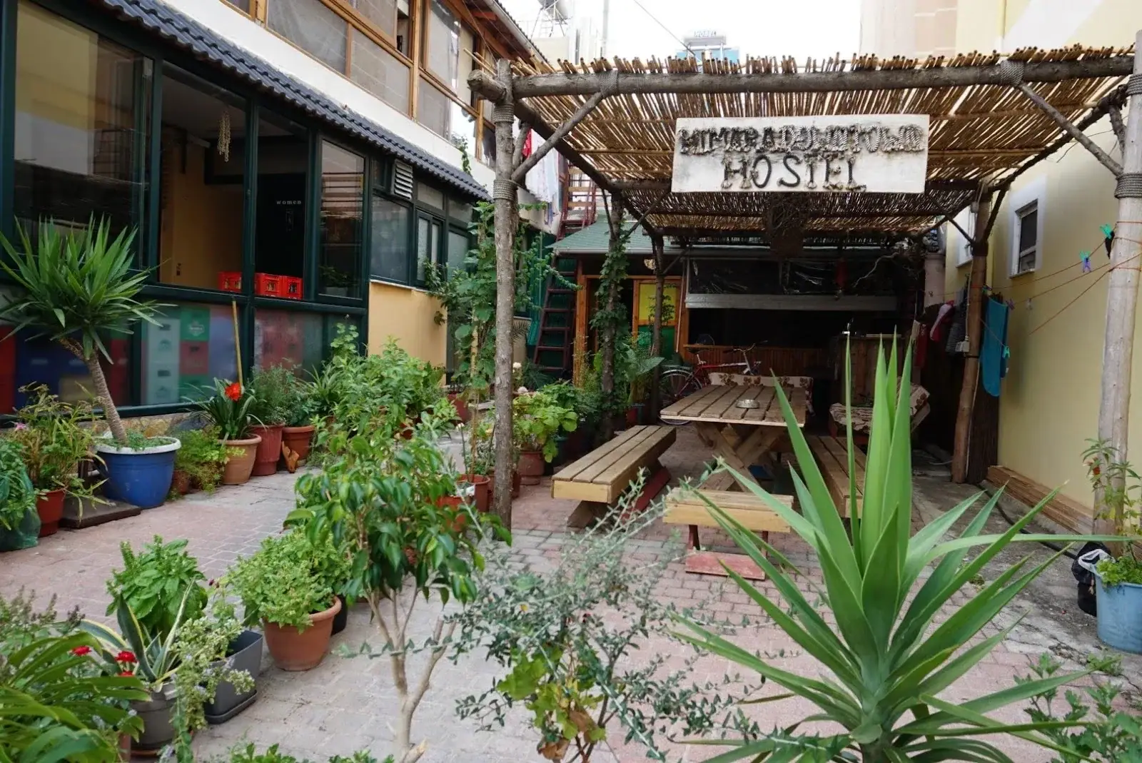 The brick courtyard at Himara Downtown Hostel, with a reed pergola carrying the hostel's painted sign, potted plants and a long picnic table