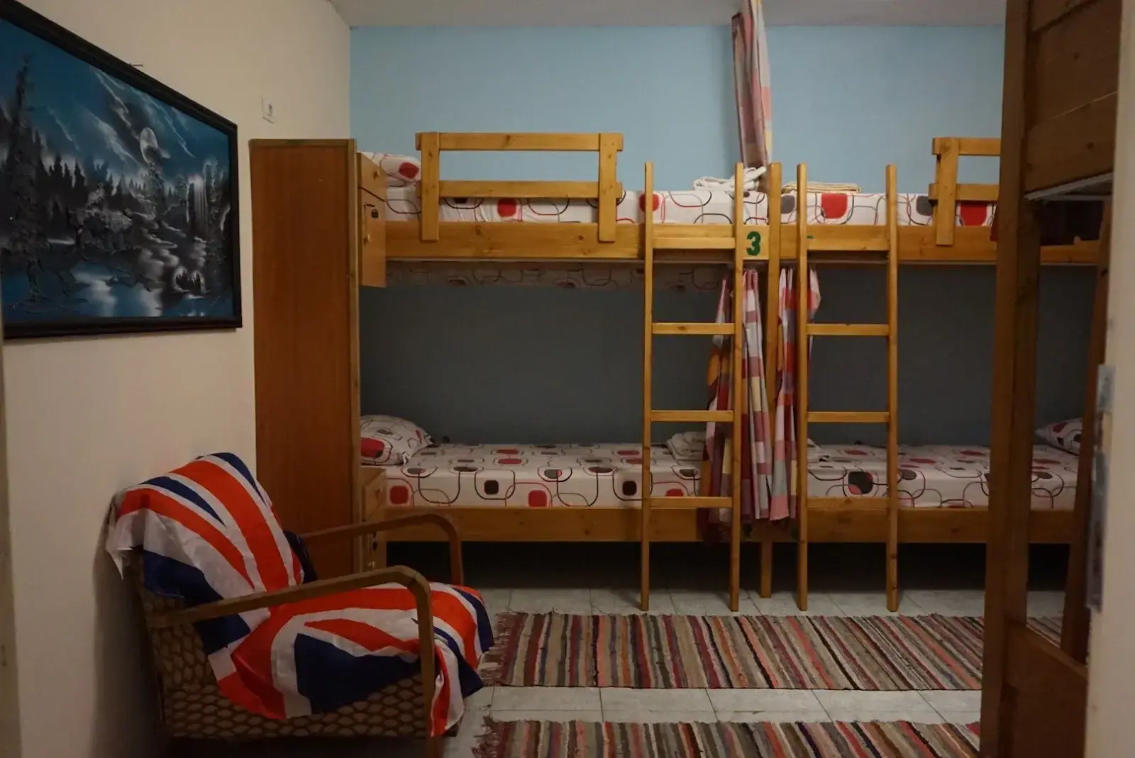 A dormitory at Himara Downtown Hostel with pine bunks, a striped rug and a wicker armchair under a painted landscape