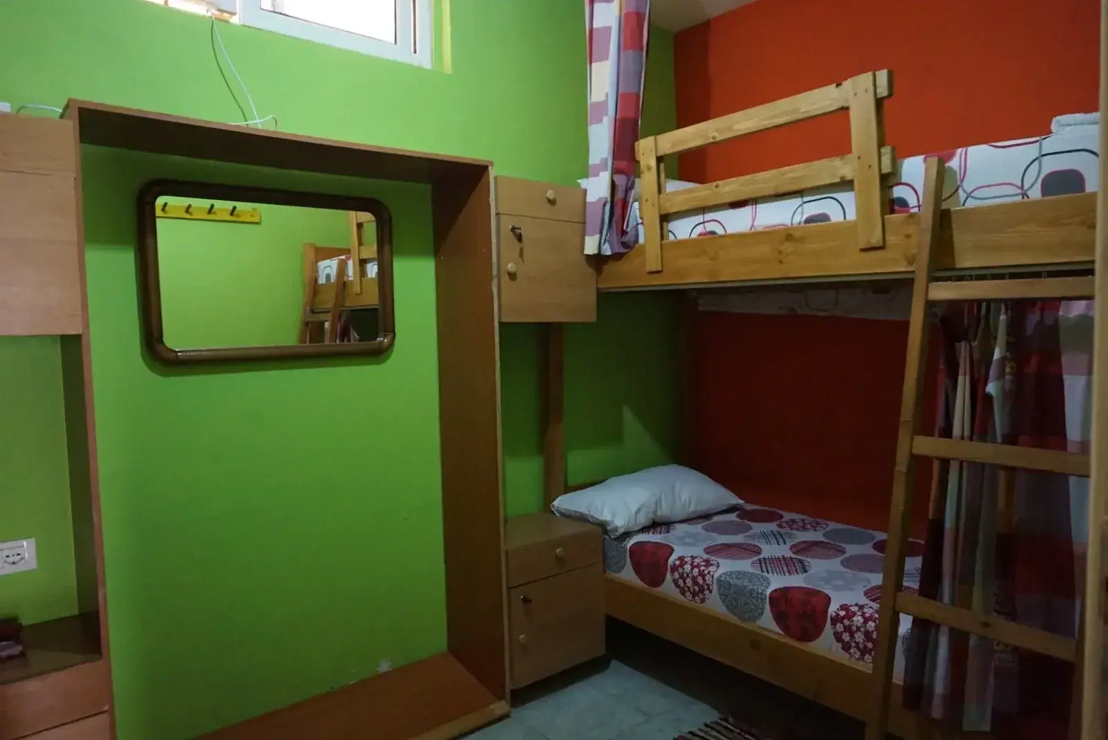 A dormitory at Himara Downtown Hostel painted green and orange, with a pine bunk, a bedside cabinet and a mirror