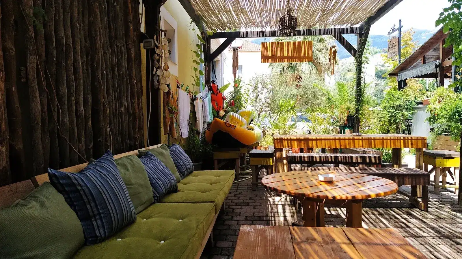 A green cushioned bench and round timber tables under a reed roof on the terrace at Himara Downtown Hostel