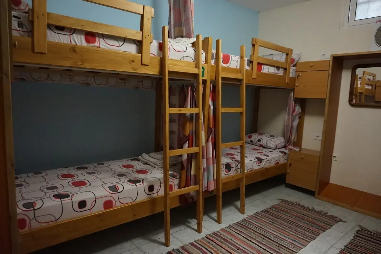 A dormitory at Himara Downtown Hostel with pine bunk beds, privacy curtains, a blue wall and a woven rug