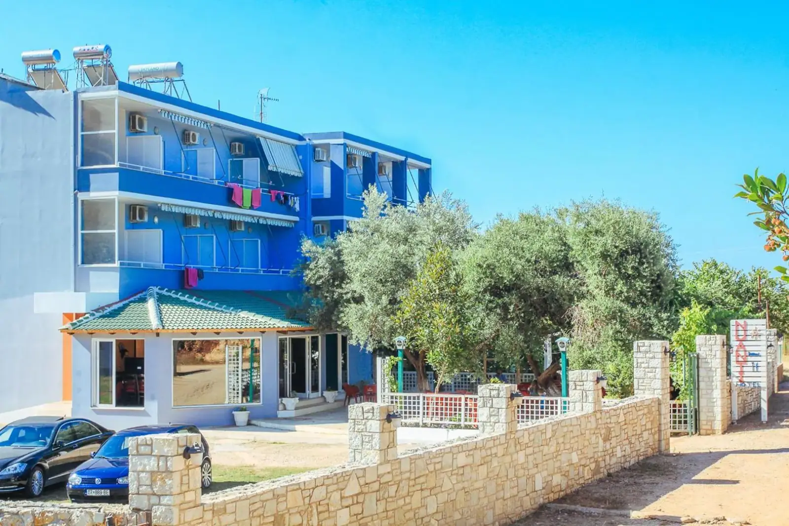 A three-storey hotel painted blue and white, standing among olive trees above a walled parking court