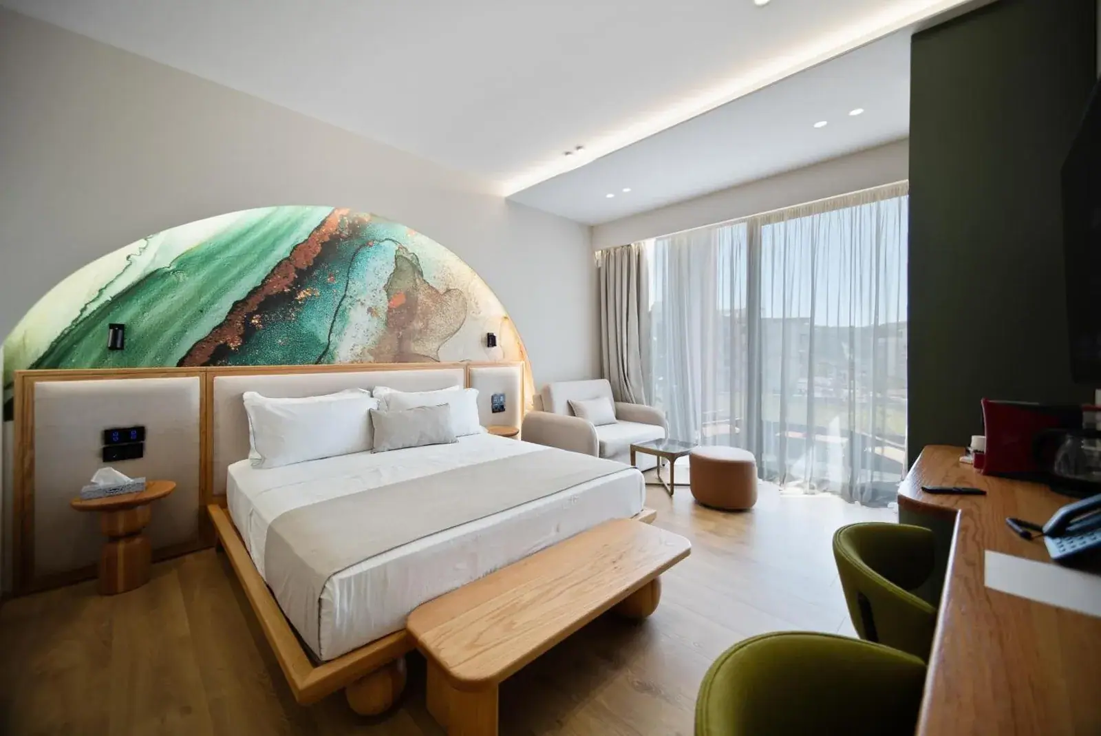 A double room at Hotel Potami with an arched painted panel in green and copper on the wall above an upholstered headboard, a pale timber floor, olive-green chairs and a glazed balcony door