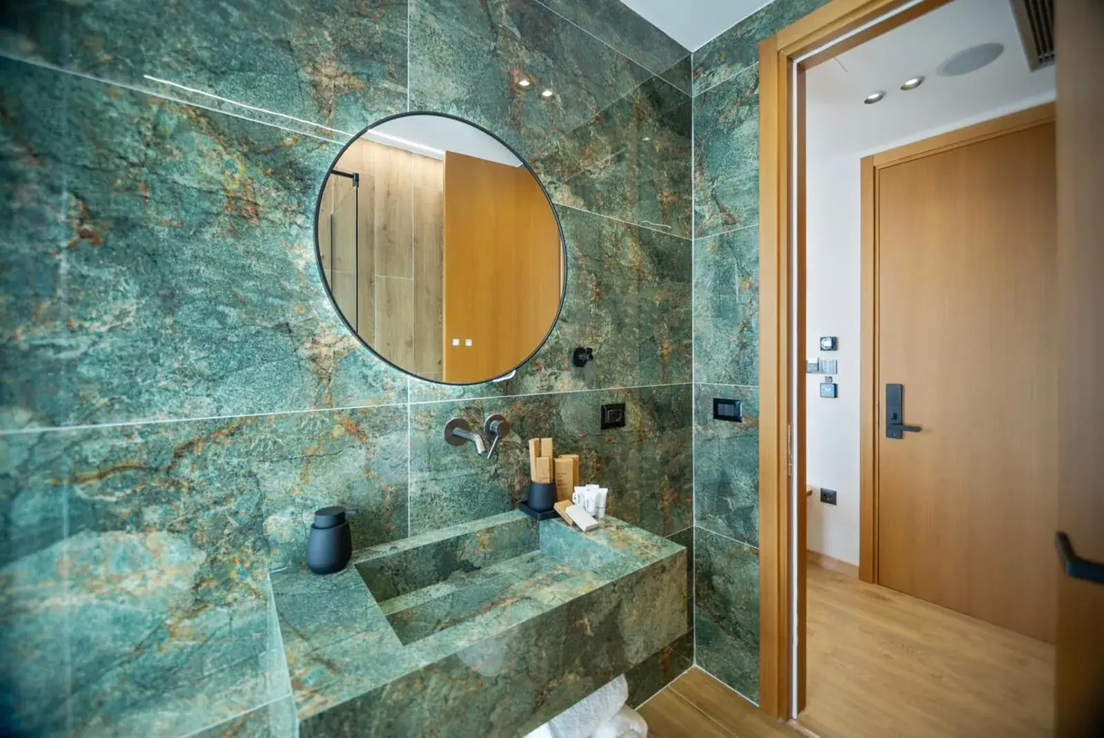 A bathroom at Hotel Potami lined in green marble, with a round black-framed mirror above a basin and counter built from mitred panels of the same green stone