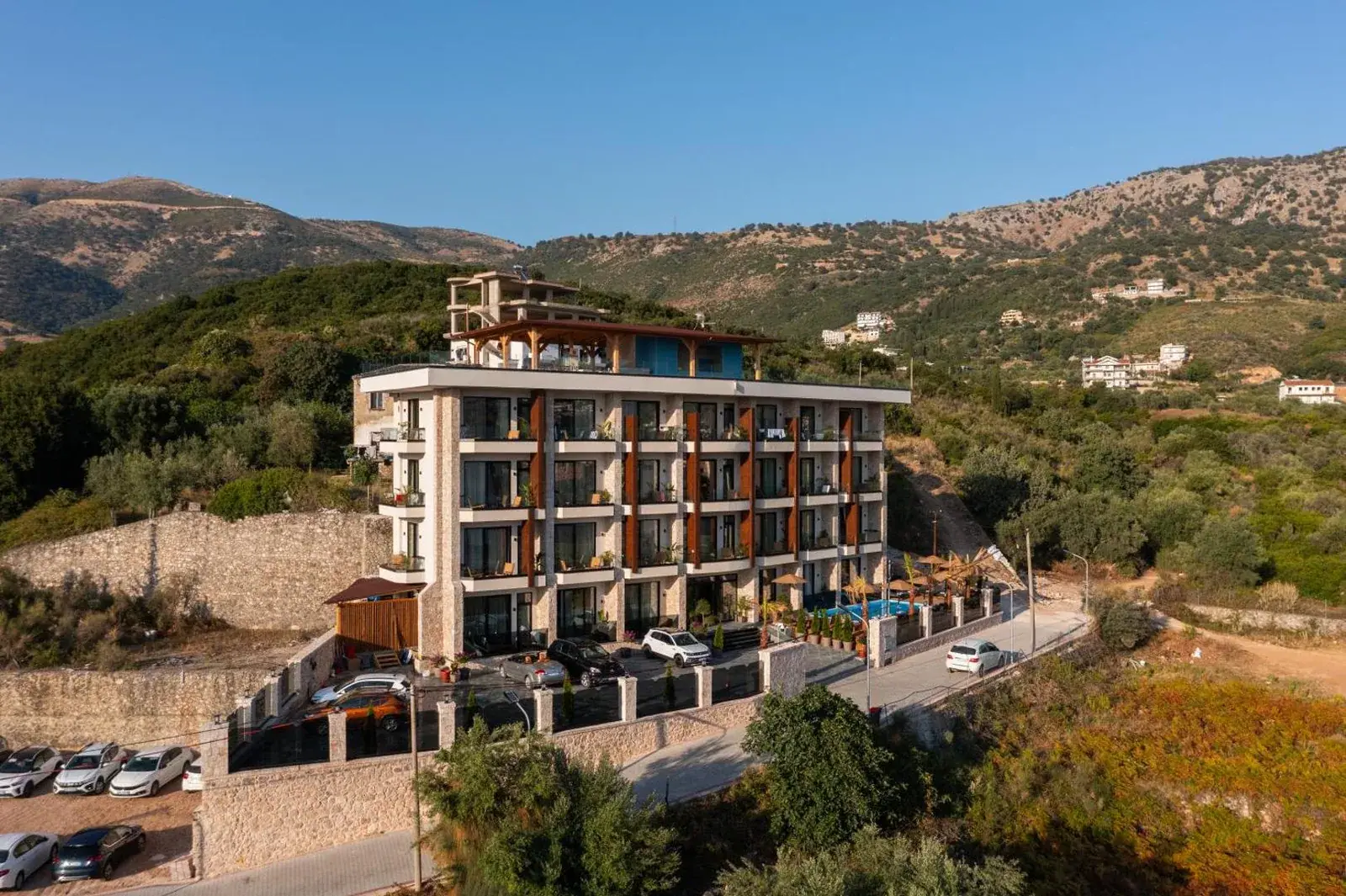 The facade of Hotel Potami: three storeys of balconies stacked above a glazed ground floor, corten fins between them, and the hills behind Himarë rising past the roof