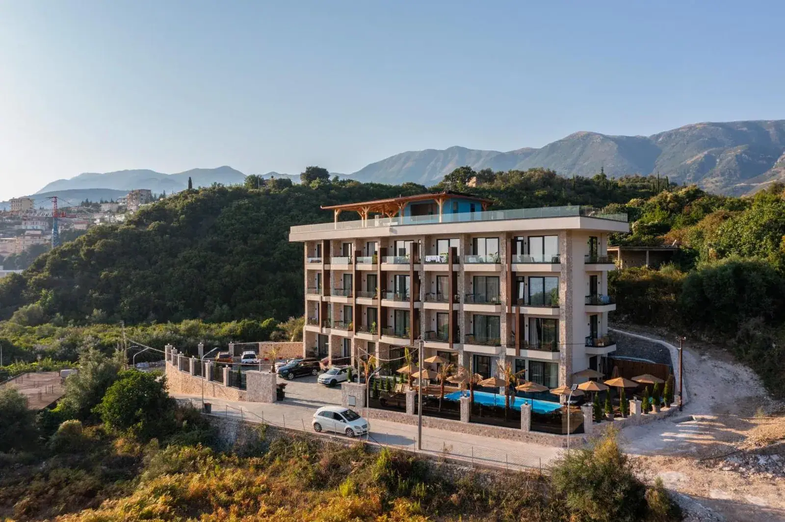 Hotel Potami seen from the air on its hillside plot above Potam, with the pool deck along the near side and the Ceraunian ridge behind