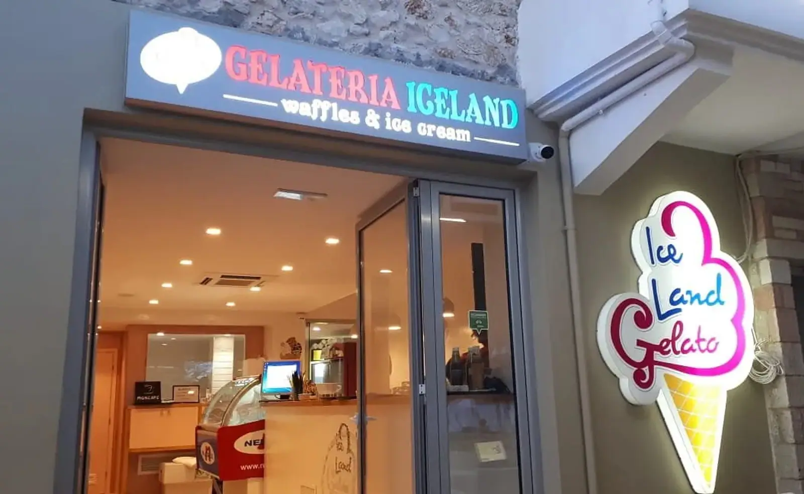 The lit shopfront of Gelateria Iceland in Spile, Himarë, its fascia reading GELATERIA ICELAND above an open bi-fold door