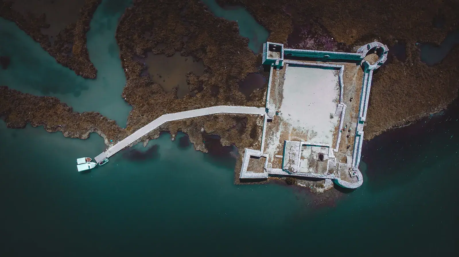 Aerial view of Ali Pasha's fortress at the mouth of the Vivari Channel: a walled rectangle with round corner towers, reached by a boardwalk across the marsh