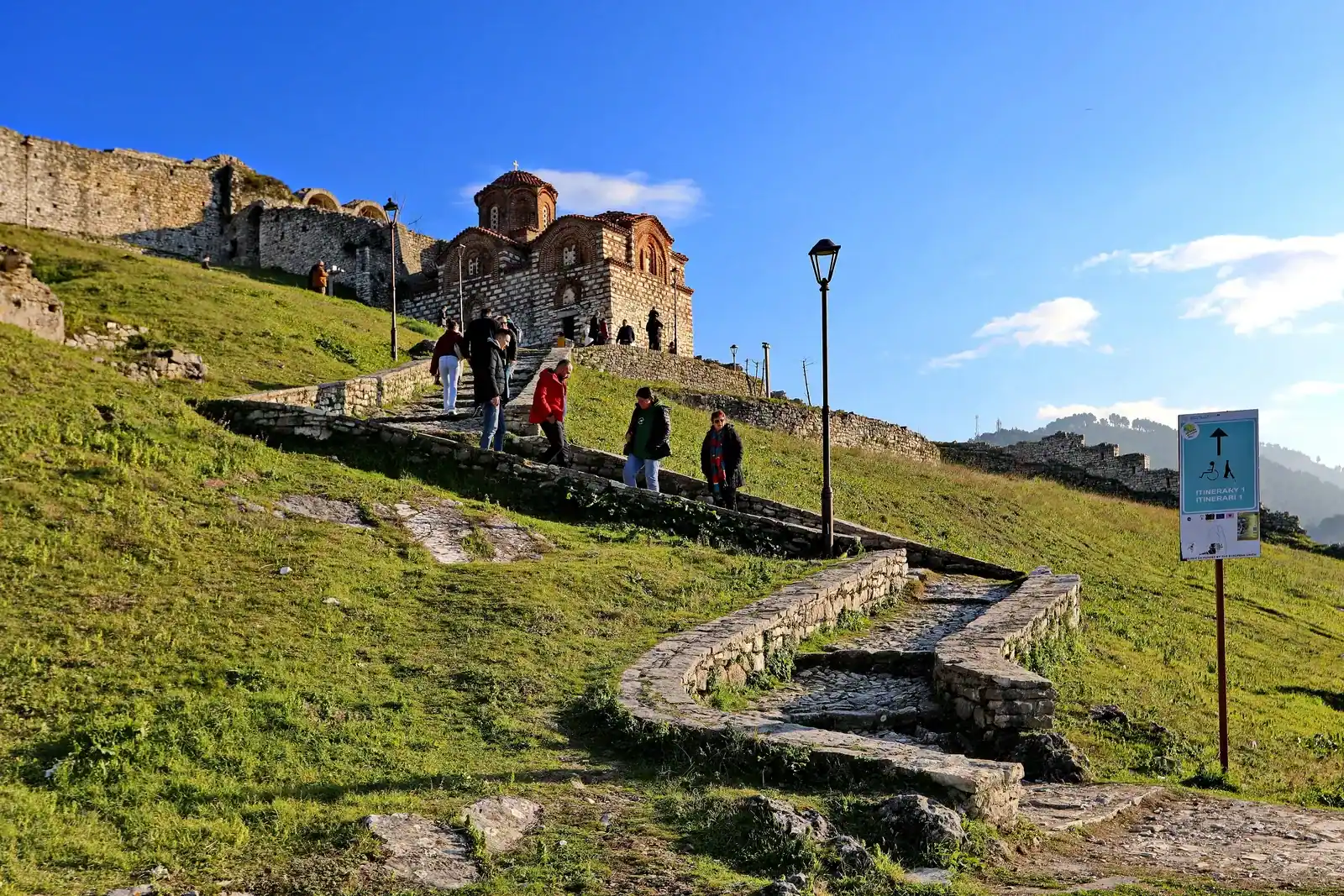 Visitors climbing a stone stairway across a grassy slope to the domed Church of the Holy Trinity, with Berat Castle's walls along the ridge behind