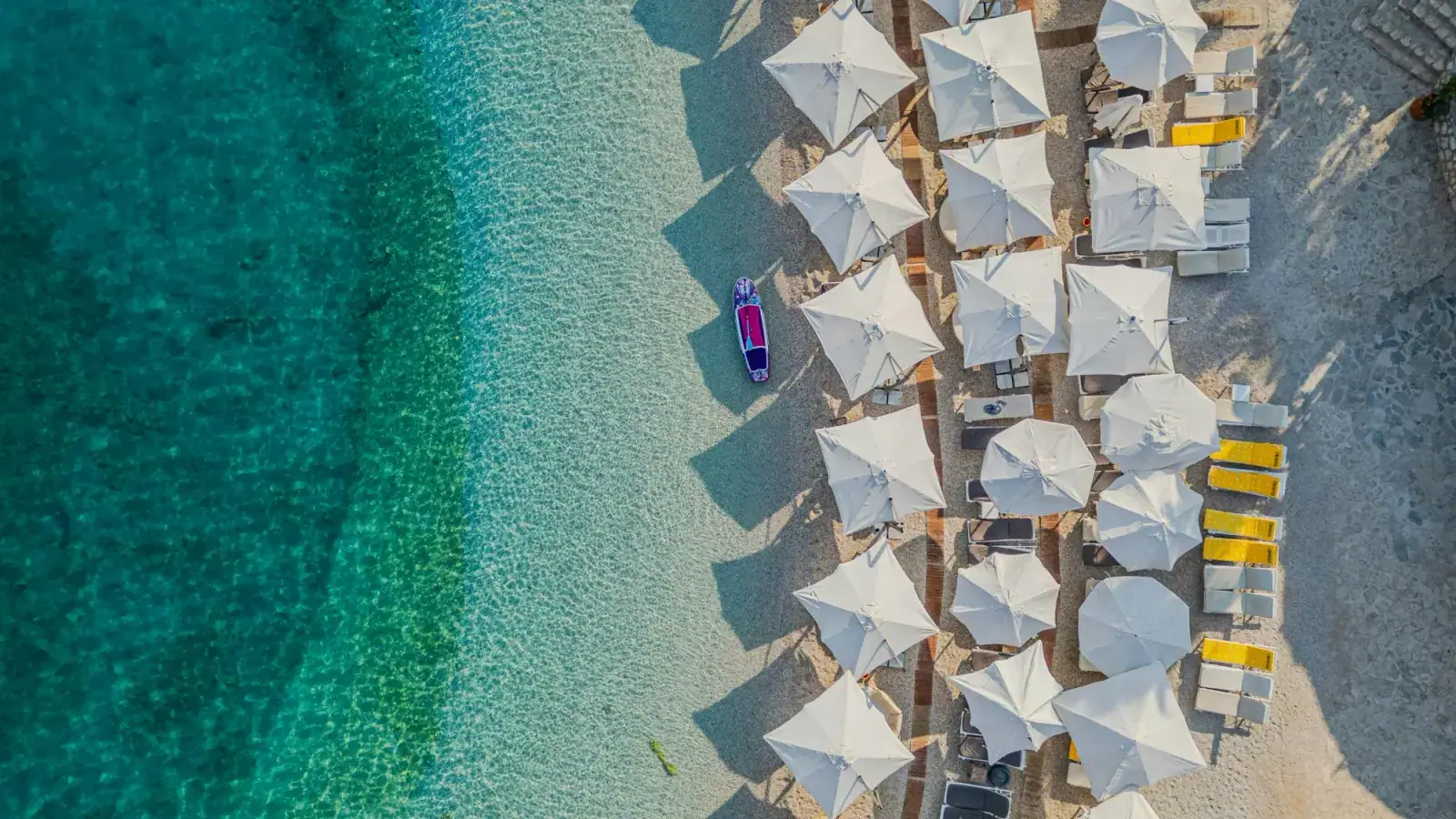 Aerial view of Dhermi Beach with rows of white sunbed umbrellas and a SUP board on turquoise Ionian water