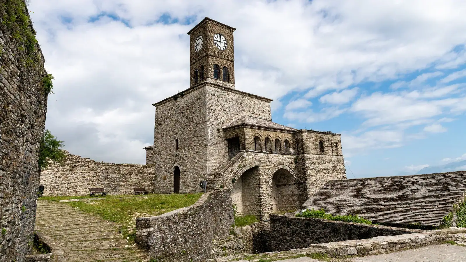 Round stone bastion topped by a clock tower and long grey fortress walls flying flags, rising above green slopes and a slate-roofed house