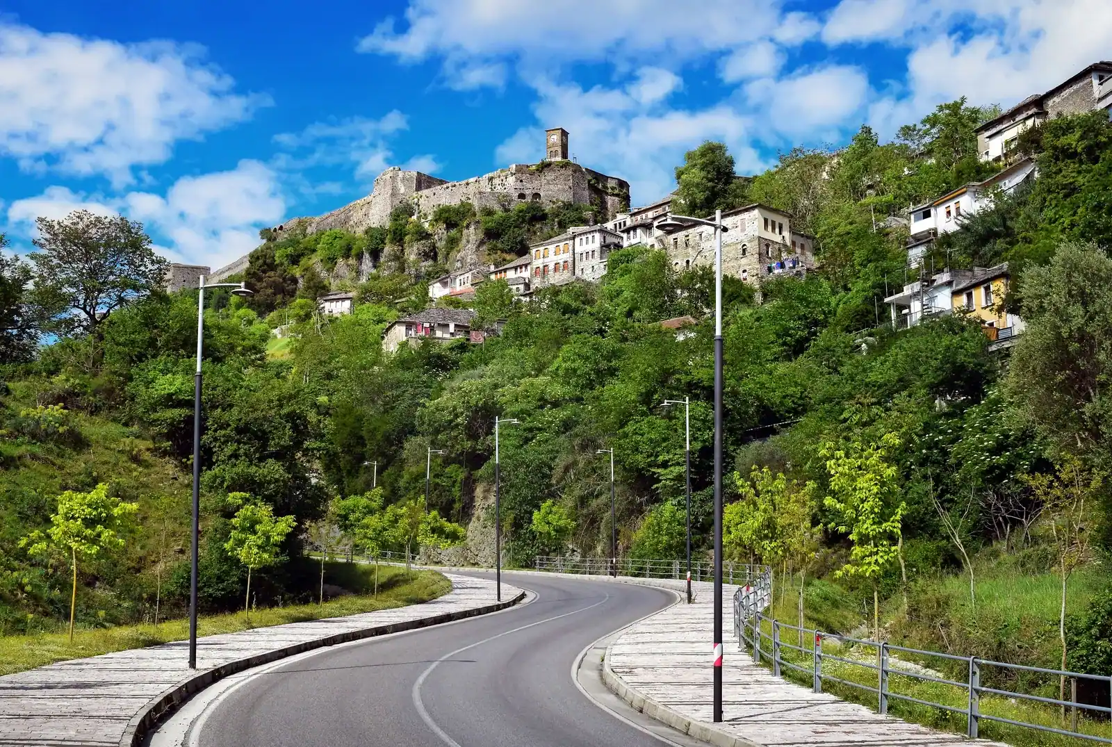 Curving road lined with street lamps leading up toward Gjirokastër, stone houses on a wooded slope and the castle with its clock tower on the crest