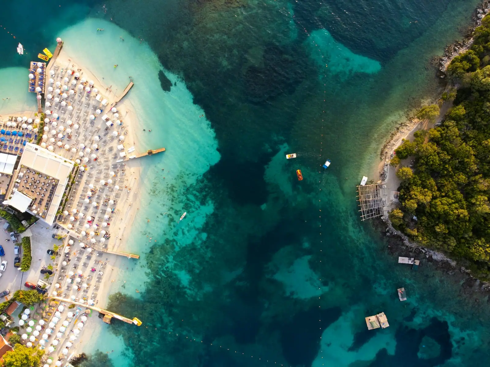 Aerial of a Ksamil beach club with white umbrellas facing a wooded islet across turquoise water