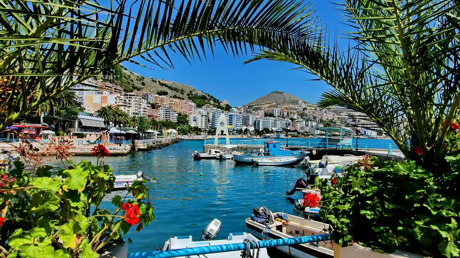 Small boats moored in Saranda's harbour framed by palm fronds, with the seafront apartment blocks and hills behind