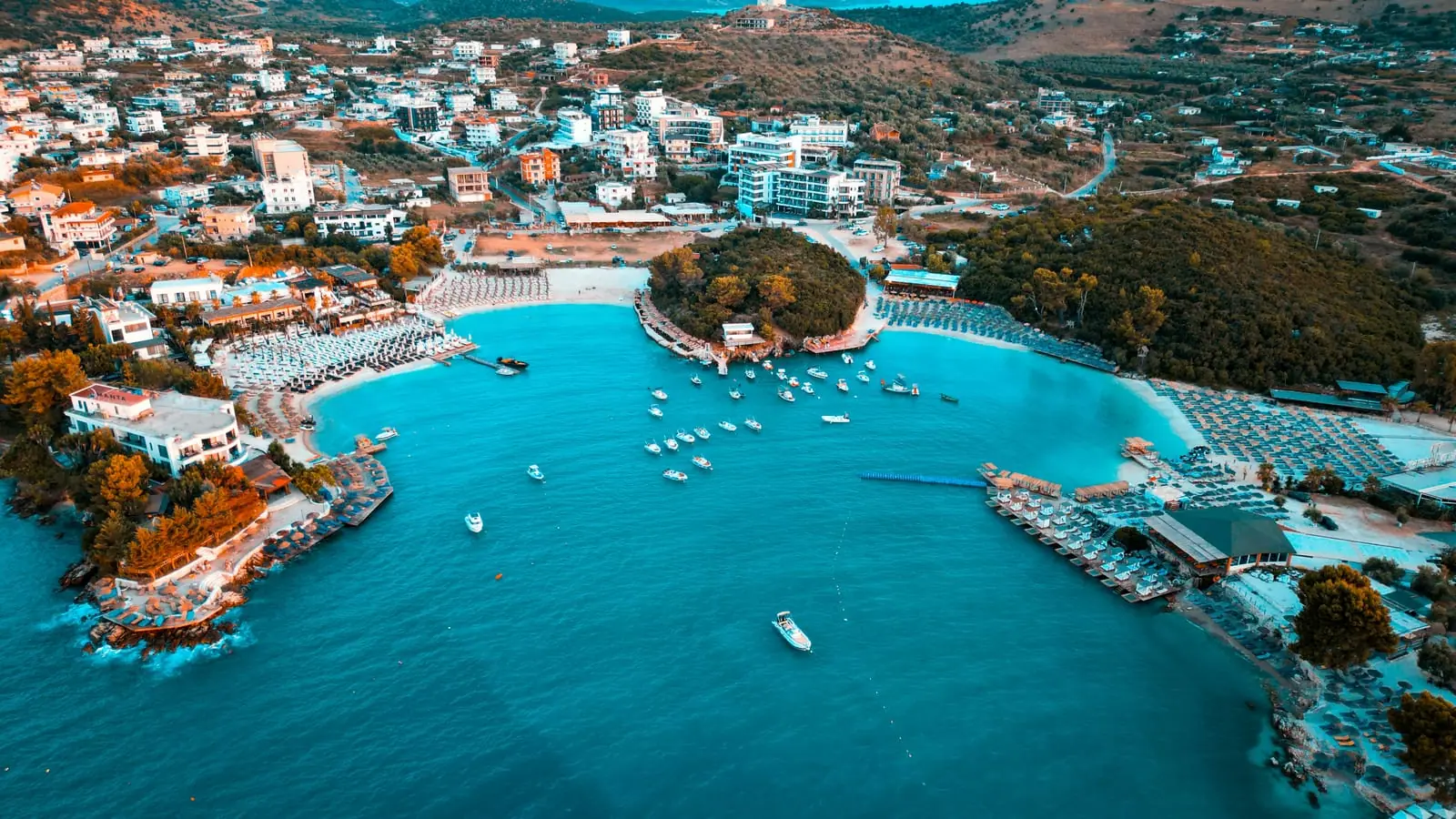 Aerial of Ksamil town and beach clubs at golden hour, the resort bay where most hotels and apartments cluster