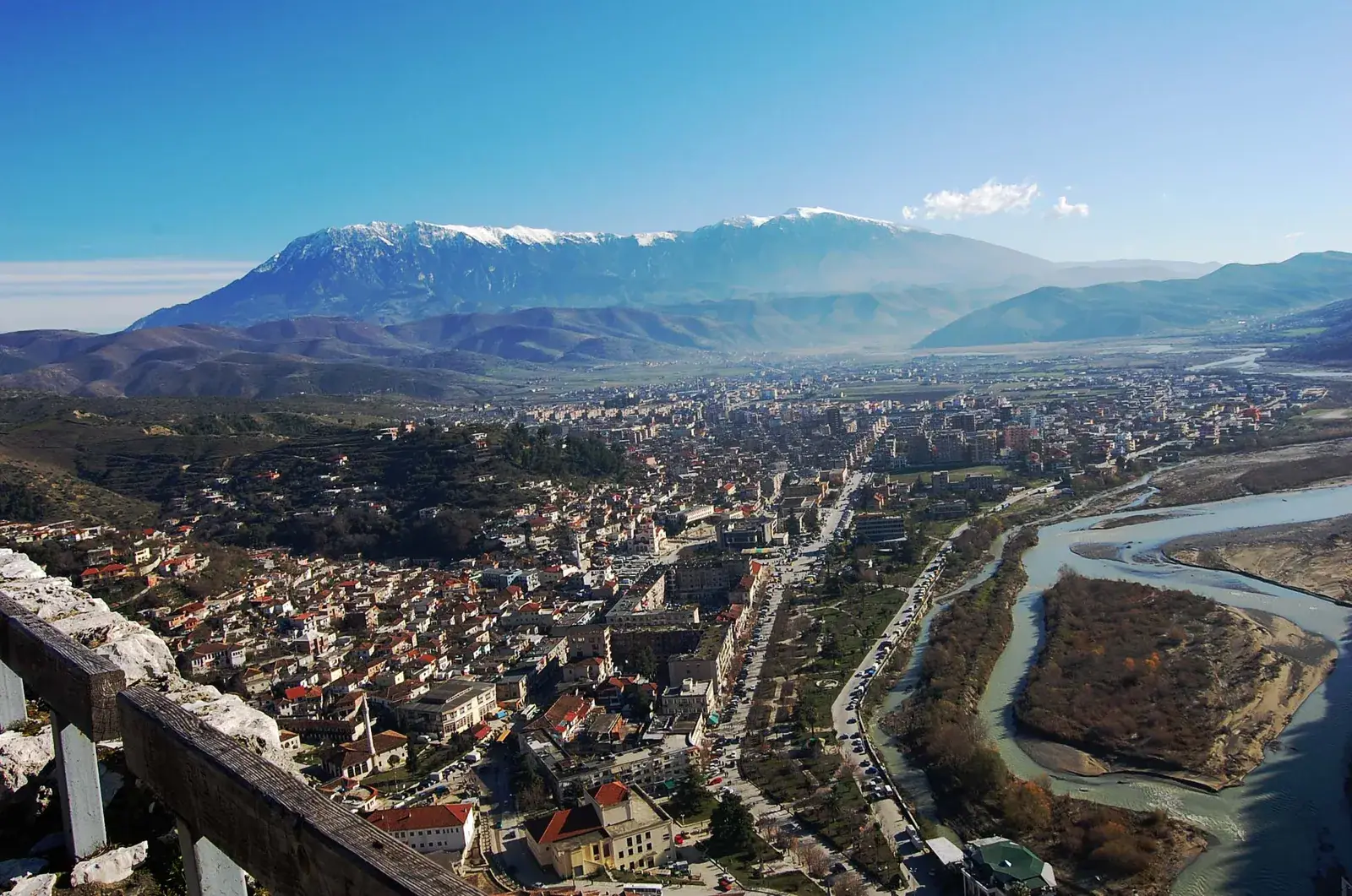 Aerial-style view from a castle railing over Berat's rooftops and the braided Osum river toward the snow-capped ridge of Mount Tomorr