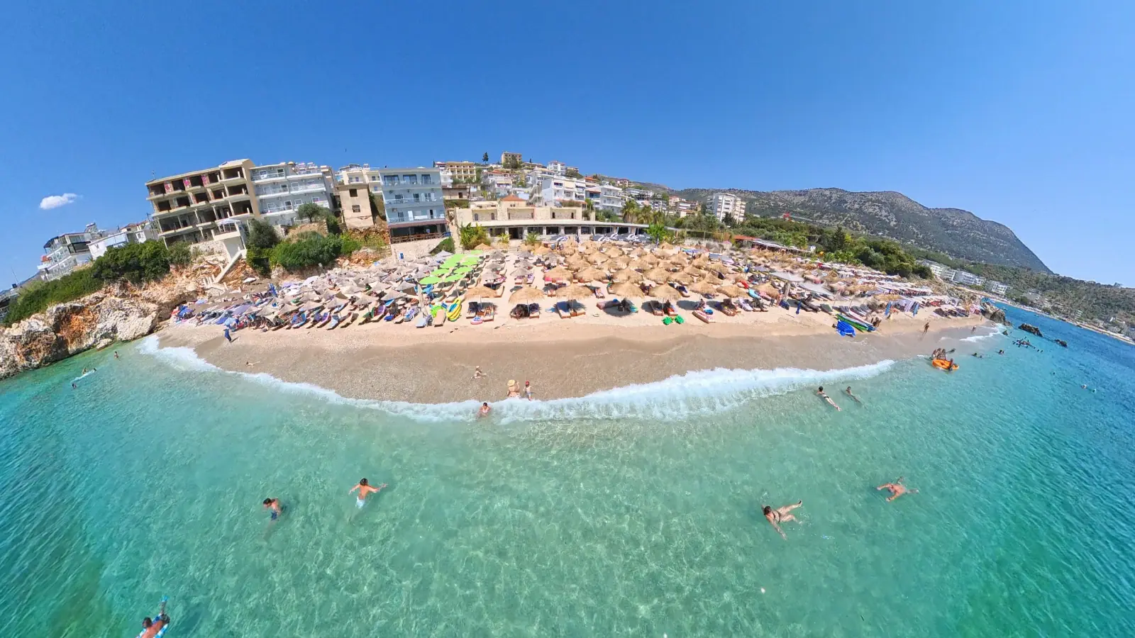 Maraçi Beach from the air, thatched umbrellas on a pebble cove below Himara's hillside hotels