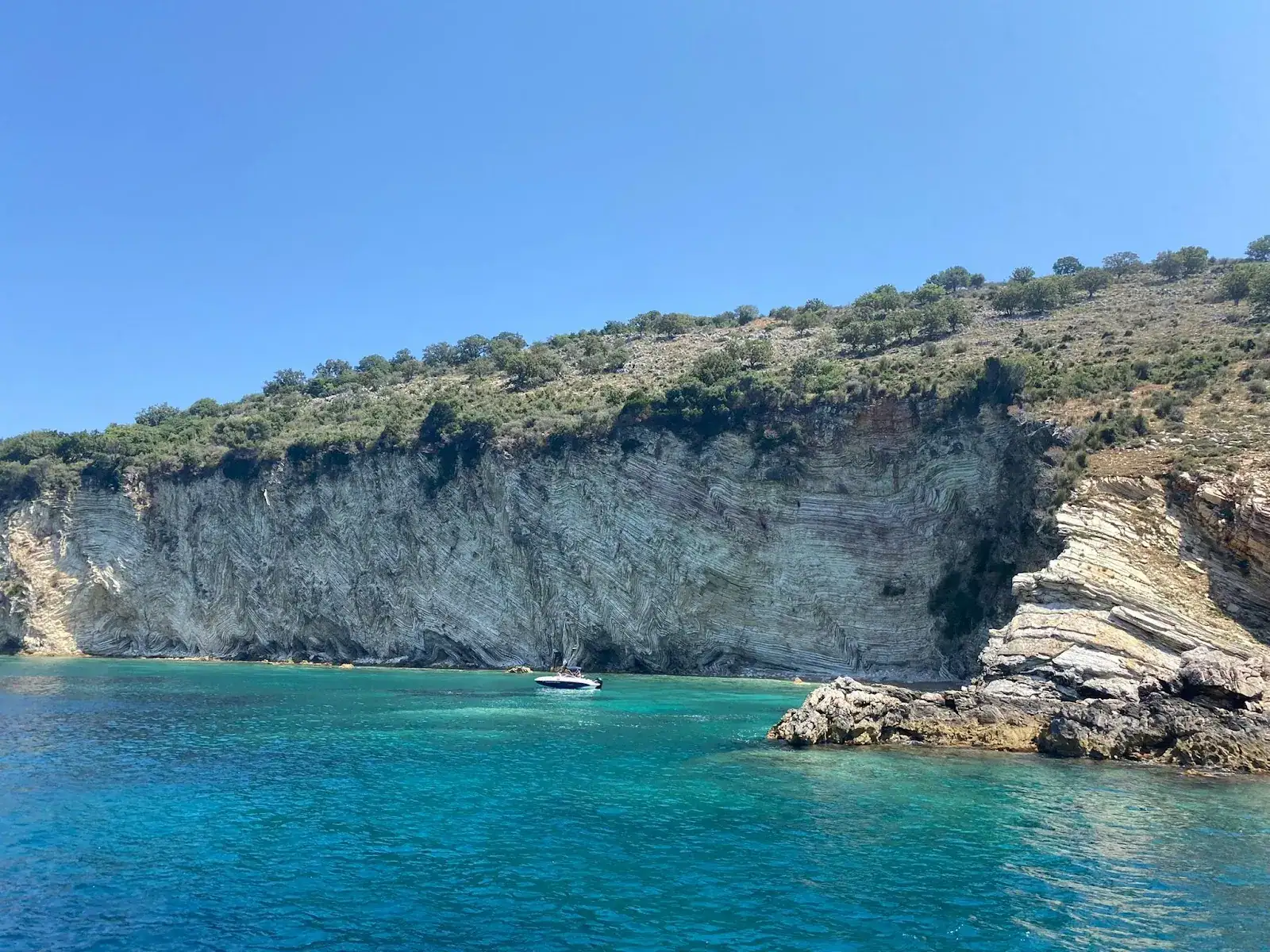 Folded grey limestone cliff at the Cape of Qefali, its rock layers bent into tight zig-zags above turquoise water with a small boat anchored below