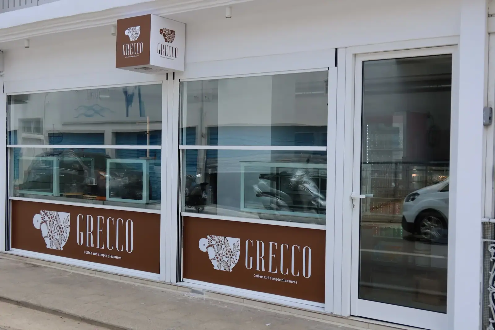 Grecco's shopfront in Himarë, glass display cases behind two windows carrying its coffee-cup logo