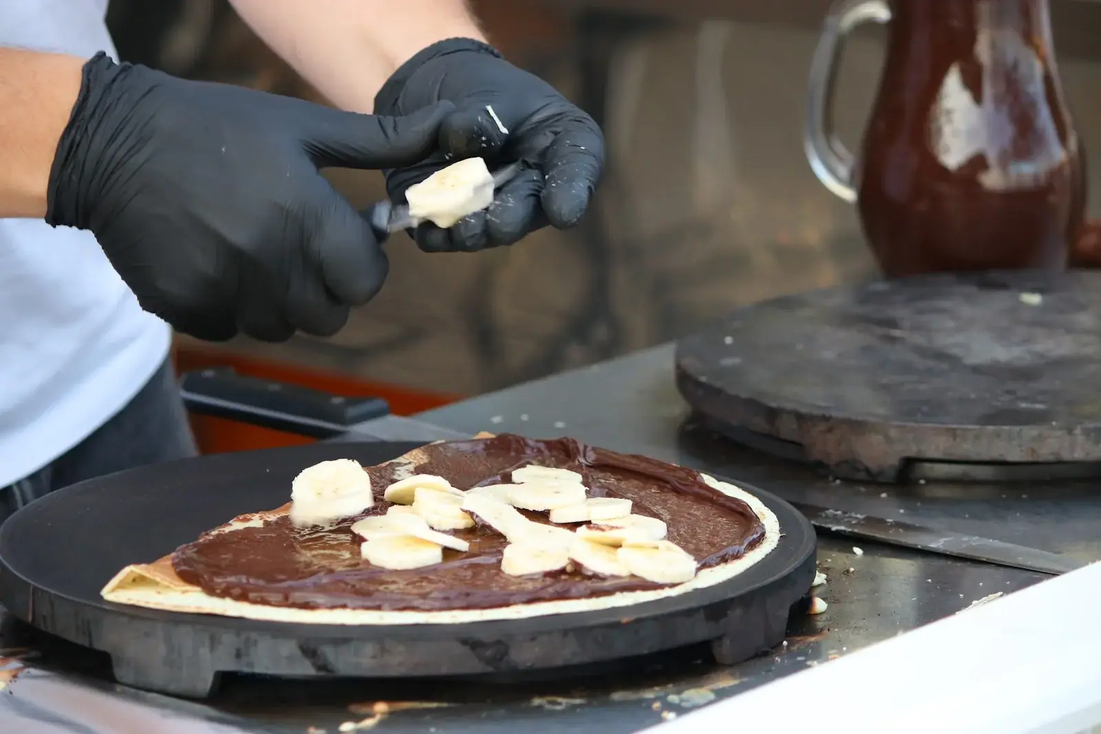 Gloved hands making a Nutella and banana crepe on the griddle at Happy Crepes in Himara