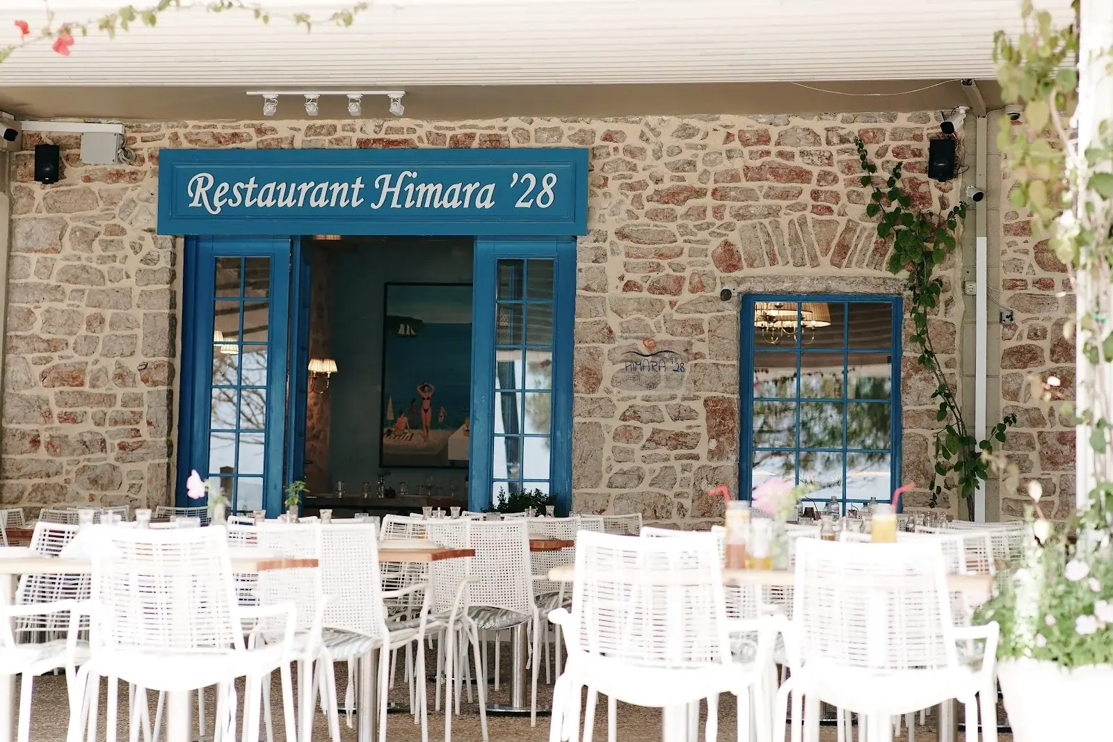 Blue-framed stone facade with the Restaurant Himara '28 sign above its terrace seating